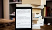 E-reader with text displayed next to a stack of physical books on a wooden table.
