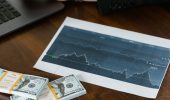 A desk scene with cash bundles, a chart printout, and a person working on a laptop.
