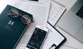 A stylish workspace featuring financial documents, eyeglasses, an iPhone displaying stock data, and a laptop.